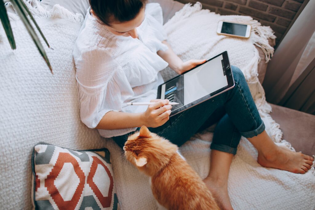 Frau sitzt mit Tablet und Ingwerkatze auf dem Sofa